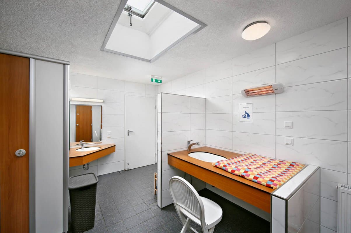 Sanitäranlagen Babybad Ein modernes Badezimmer mit einem hölzernen Wickeltisch, einem weißen Stuhl, einem Spiegel, einem Waschbecken und einer Dachluke. Wände und Boden sind in Weiß und Grau gefliest.