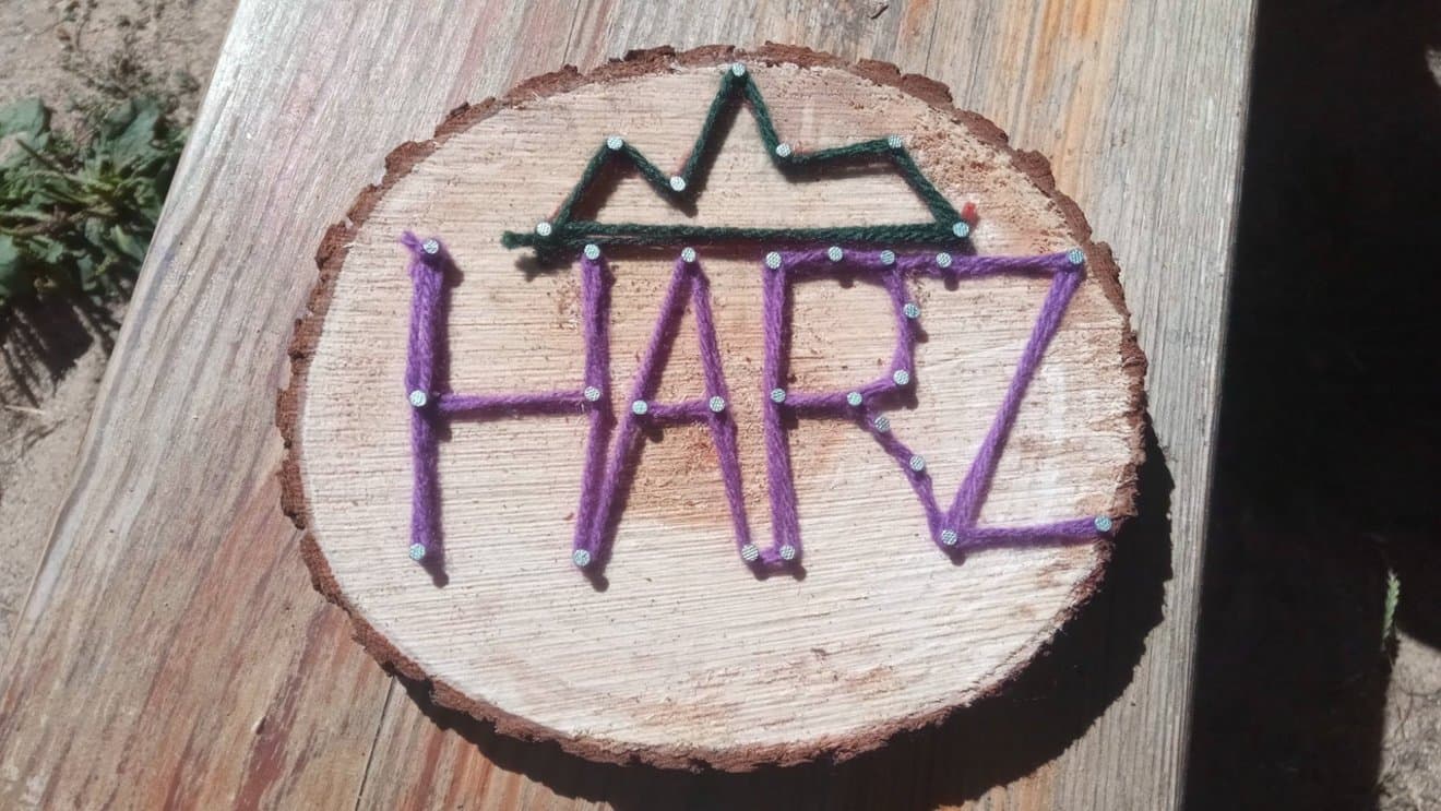 Kinderanimation Holz Holzscheibe mit „HARZ“ in lila Garn, darüber Bergform in grünem Garn, auf einer Holzfläche.