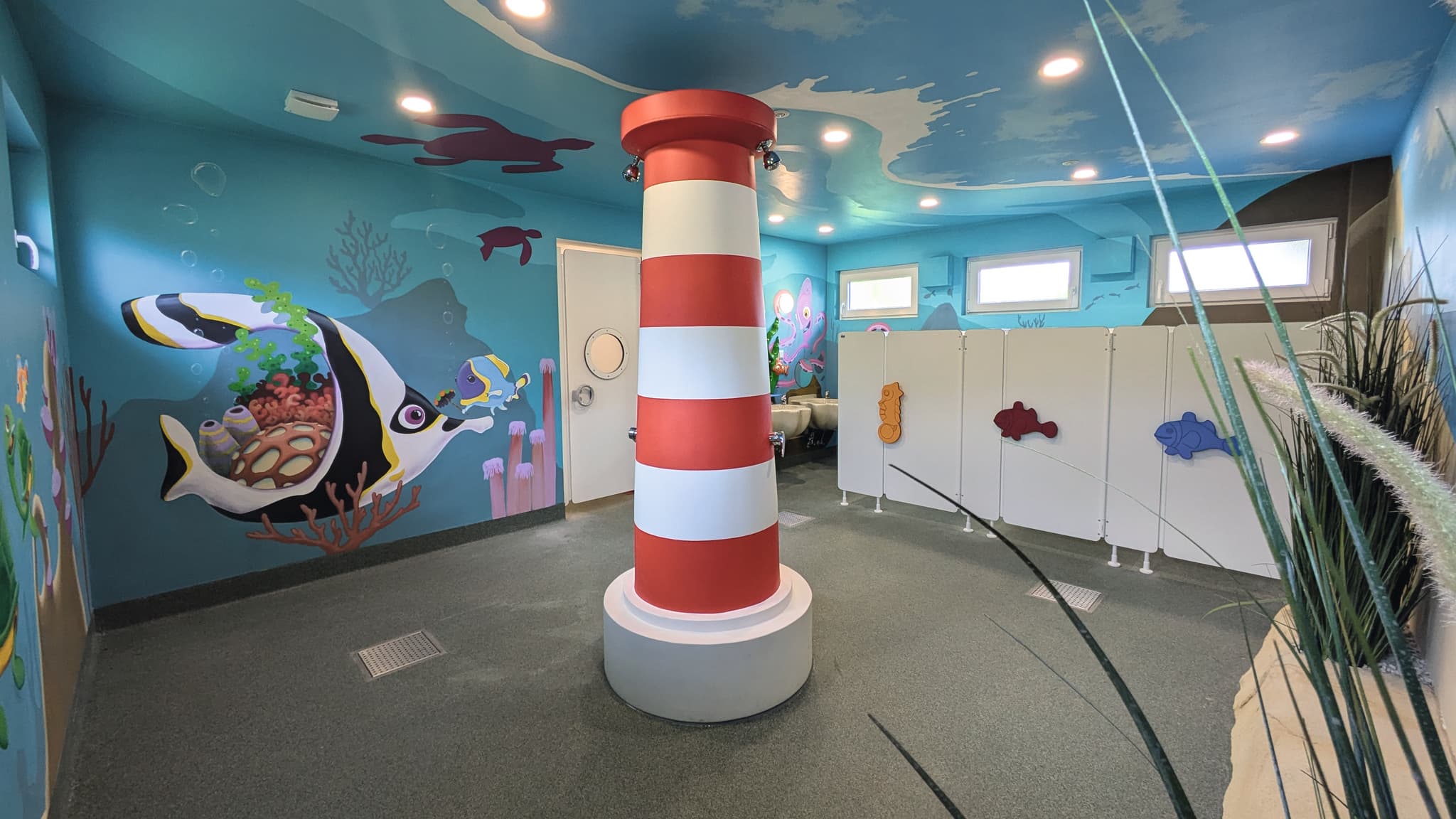 Kindererlebnisbad Farbenfrohes Kinderbad mit Wandmalereien zum Thema Meer, einer rot-weiß gestreiften Säule und Fischdekorationen an den Trennwänden.