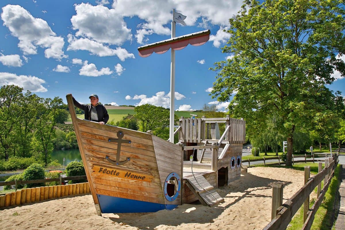 Piratenspielboot Eine Person steht auf einer hölzernen Spielschiffstruktur in einem sonnigen Park mit Bäumen und einem blauen Himmel mit Wolken.