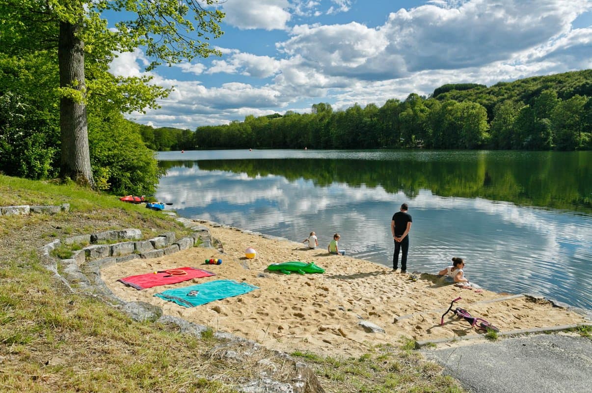 Liegewiese Sandstrand Ein ruhiger See mit einem Sandstrand, Handtüchern und Spielzeug. Die Menschen genießen das Wasser unter einem teilweise bewölkten Himmel, umgeben von üppigen grünen Bäumen.