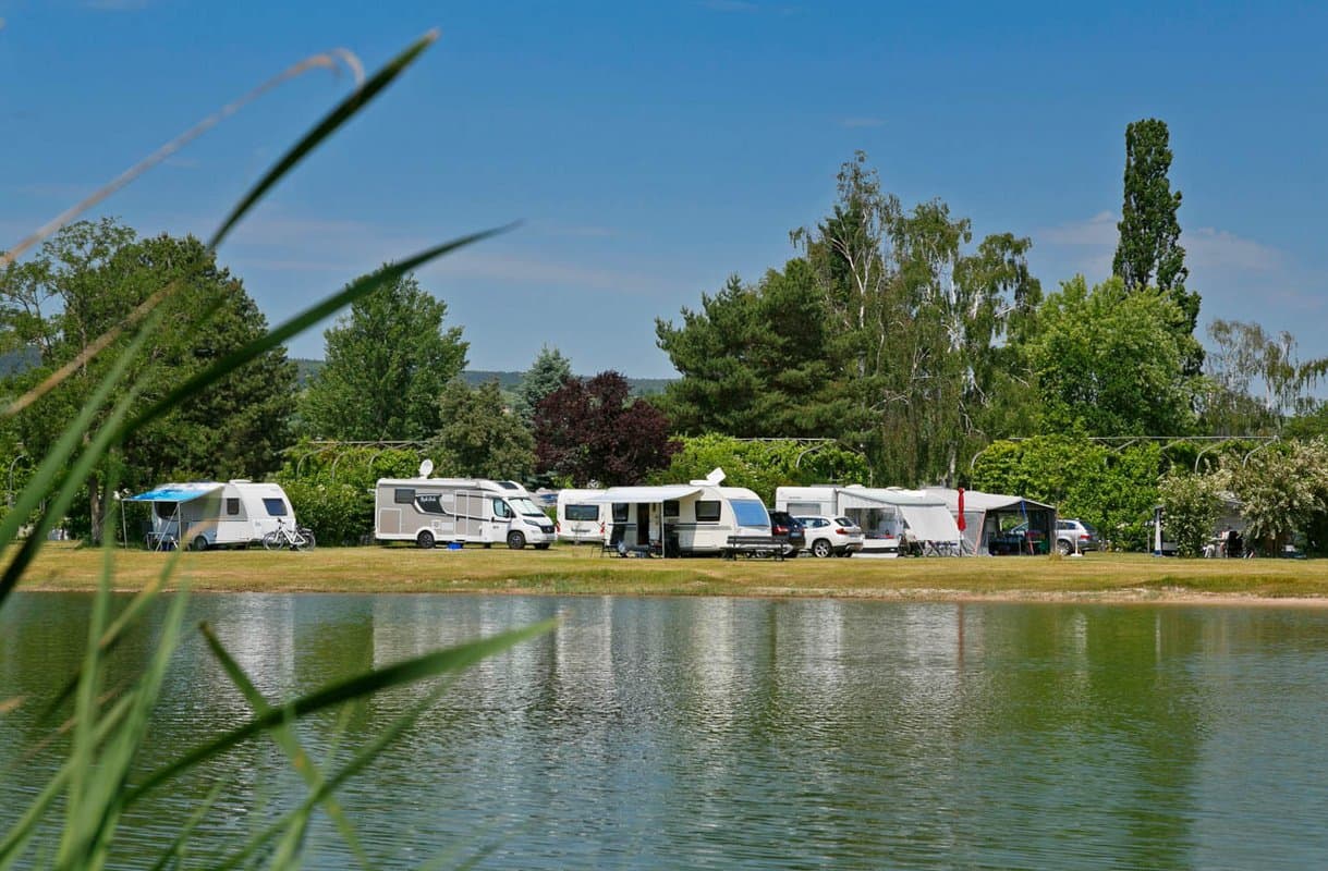 Stellplätze am Almensee Wohnwagen, die an einem ruhigen See geparkt sind, umgeben von üppigem Grün und Bäumen unter einem klaren blauen Himmel.